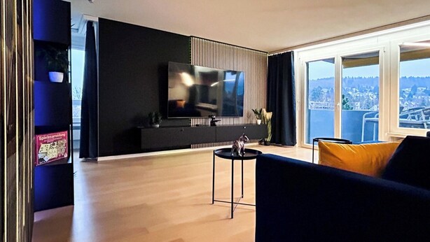 Ein modernes Wohnzimmer mit großem Fernseher, einer Couch und Blick auf eine Stadtlandschaft durch große Fenster.