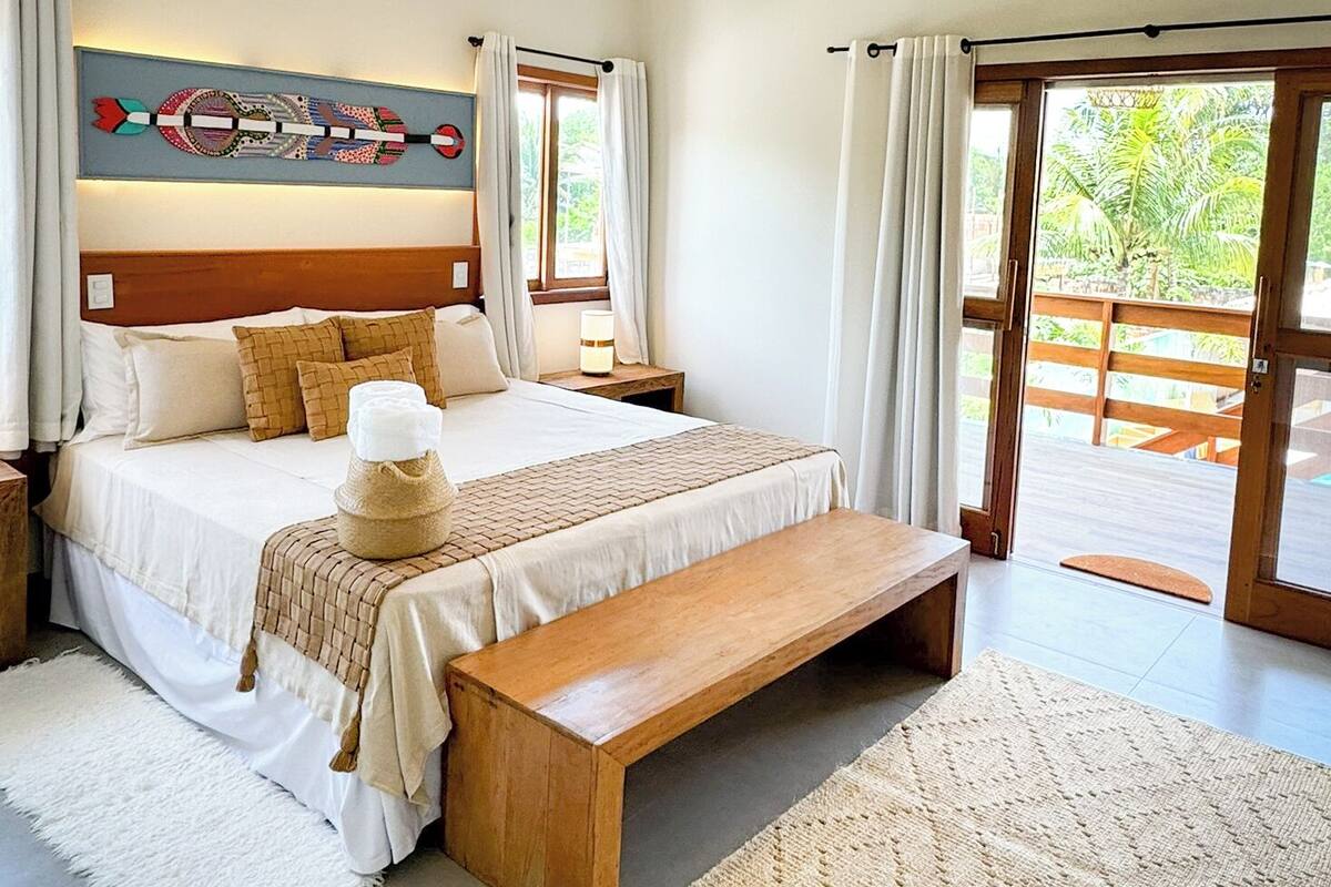 A modern bedroom with a large bed, a wooden bench, a hanging light fixture, and a view of the outdoors through a sliding door.