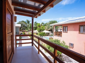 Un balcone con vista su una piscina, un edificio rosa e vegetazione circostante.