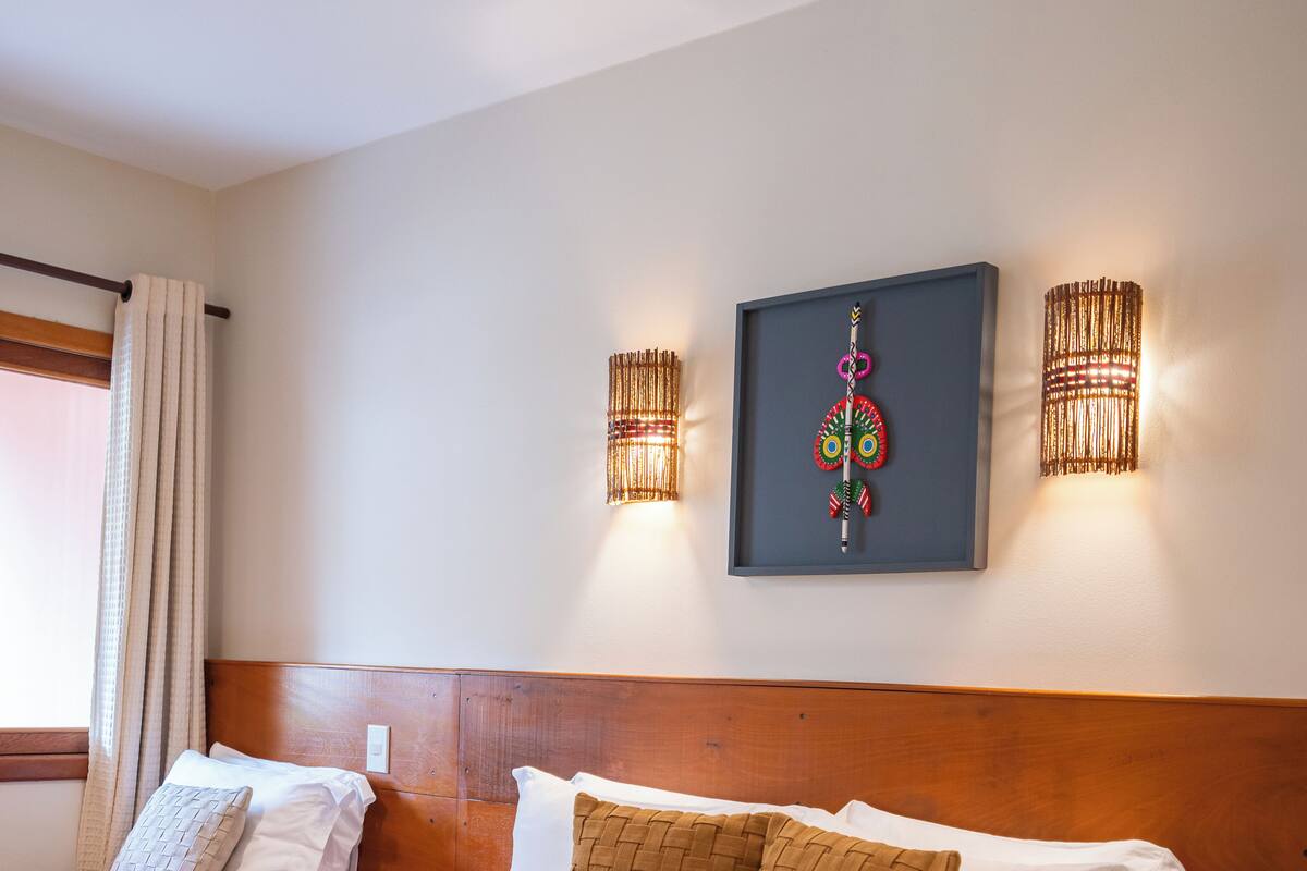A hotel room with a large bed, two bedside tables, a headboard, a window with curtains, and wall-mounted lighting.