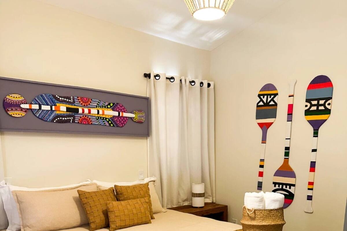 A neatly made bed with a patterned bedspread and pillows, a woven side table with a lamp, and a wall with decorative wall art and two paddle designs.
