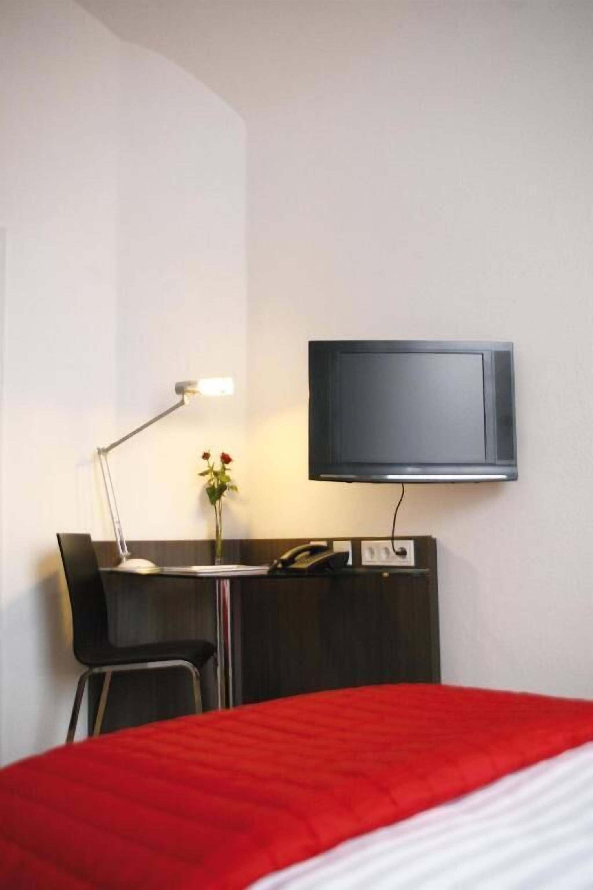 A hotel room with a bed, a desk with a chair, a desk lamp, a flat-screen TV mounted on the wall, and a vase with flowers on the desk.