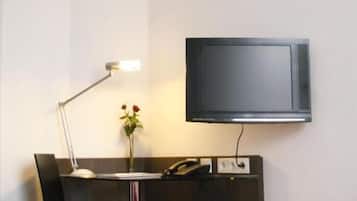 A hotel room with a bed, a desk with a chair, a desk lamp, a flat-screen TV mounted on the wall, and a vase with flowers on the desk.
