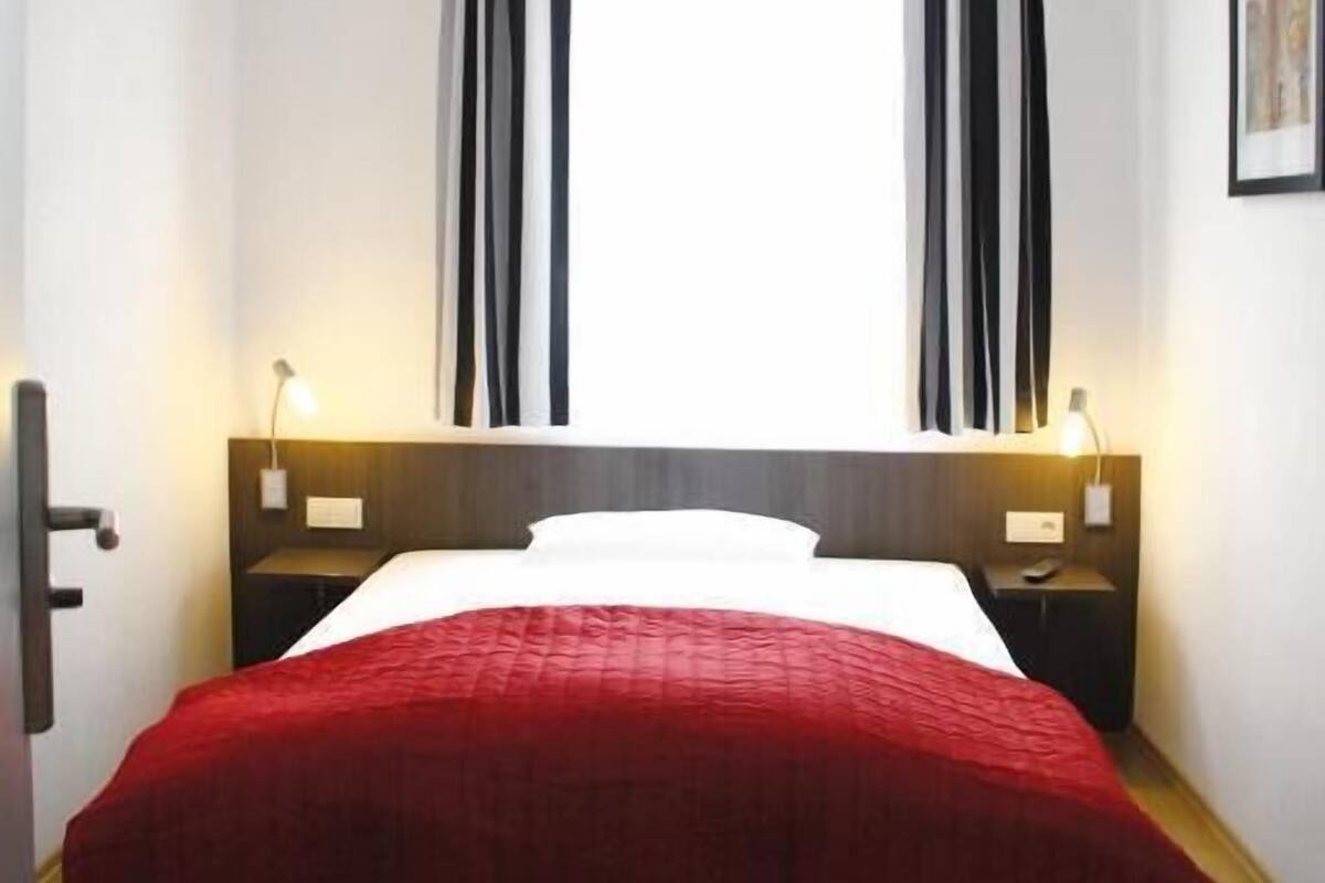 Una habitación de hotel con una cama, dos lámparas de noche, una mesita de noche y una ventana con cortinas.
