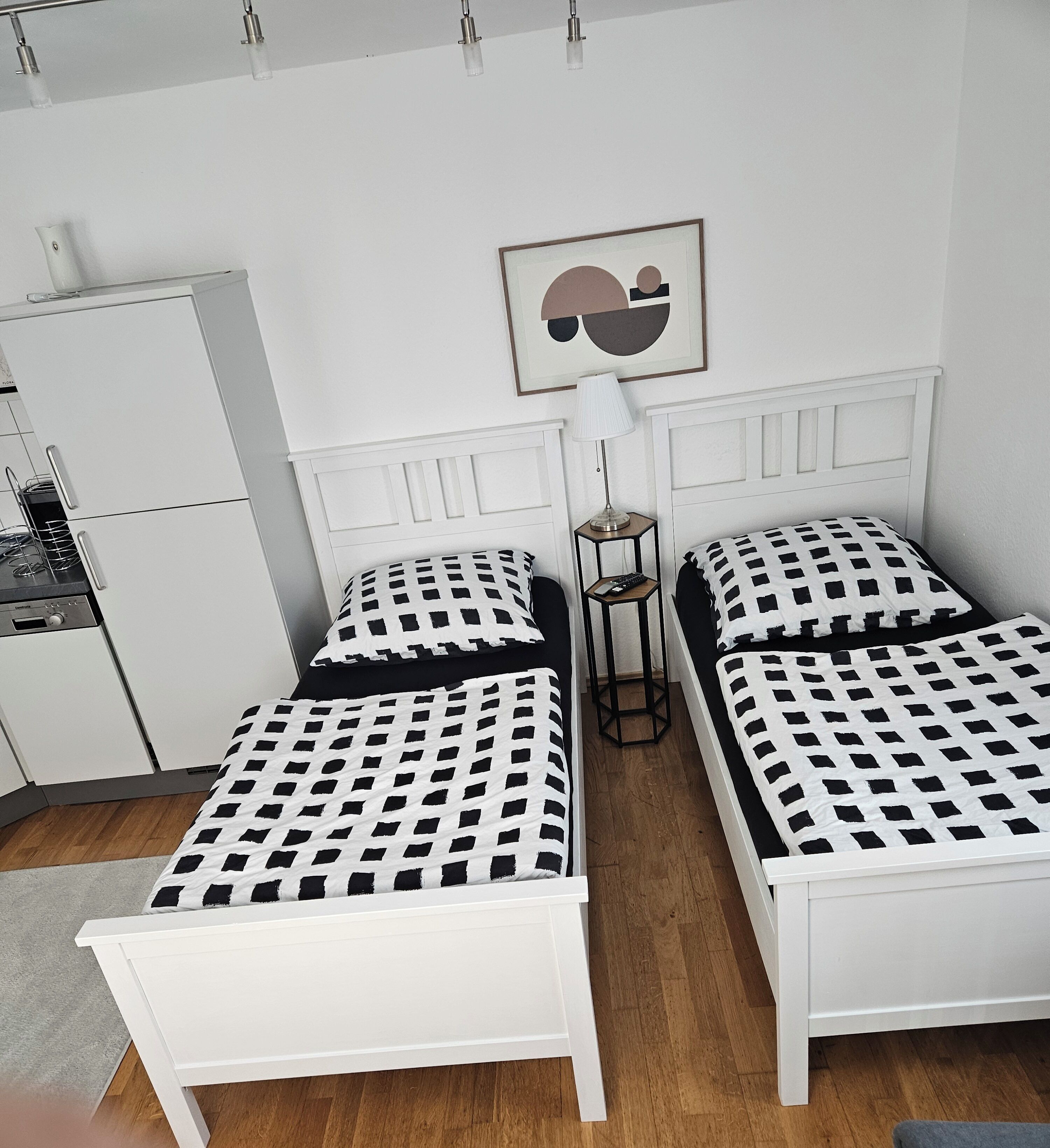 Une chambre avec deux lits blancs, recouverts d’une literie à carreaux noirs et blancs, une petite table avec une lampe et un réfrigérateur.