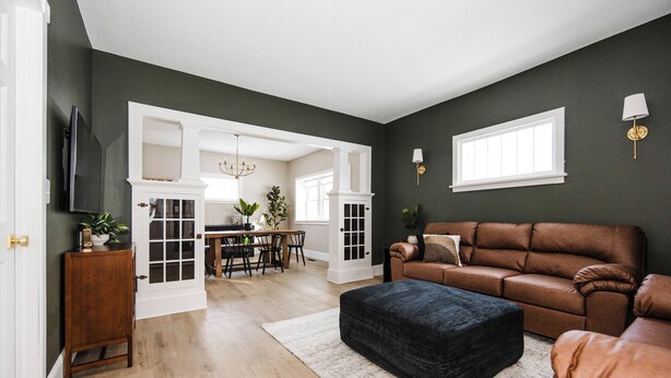 A living room with a brown leather sofa, a black ottoman, and a dining area with a chandelier.