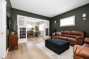 A living room with a brown leather sofa, a black ottoman, and a dining area with a chandelier. - Dog & Family Friendly, 3 Blocks to Beach & Bars (Detroit Lakes)