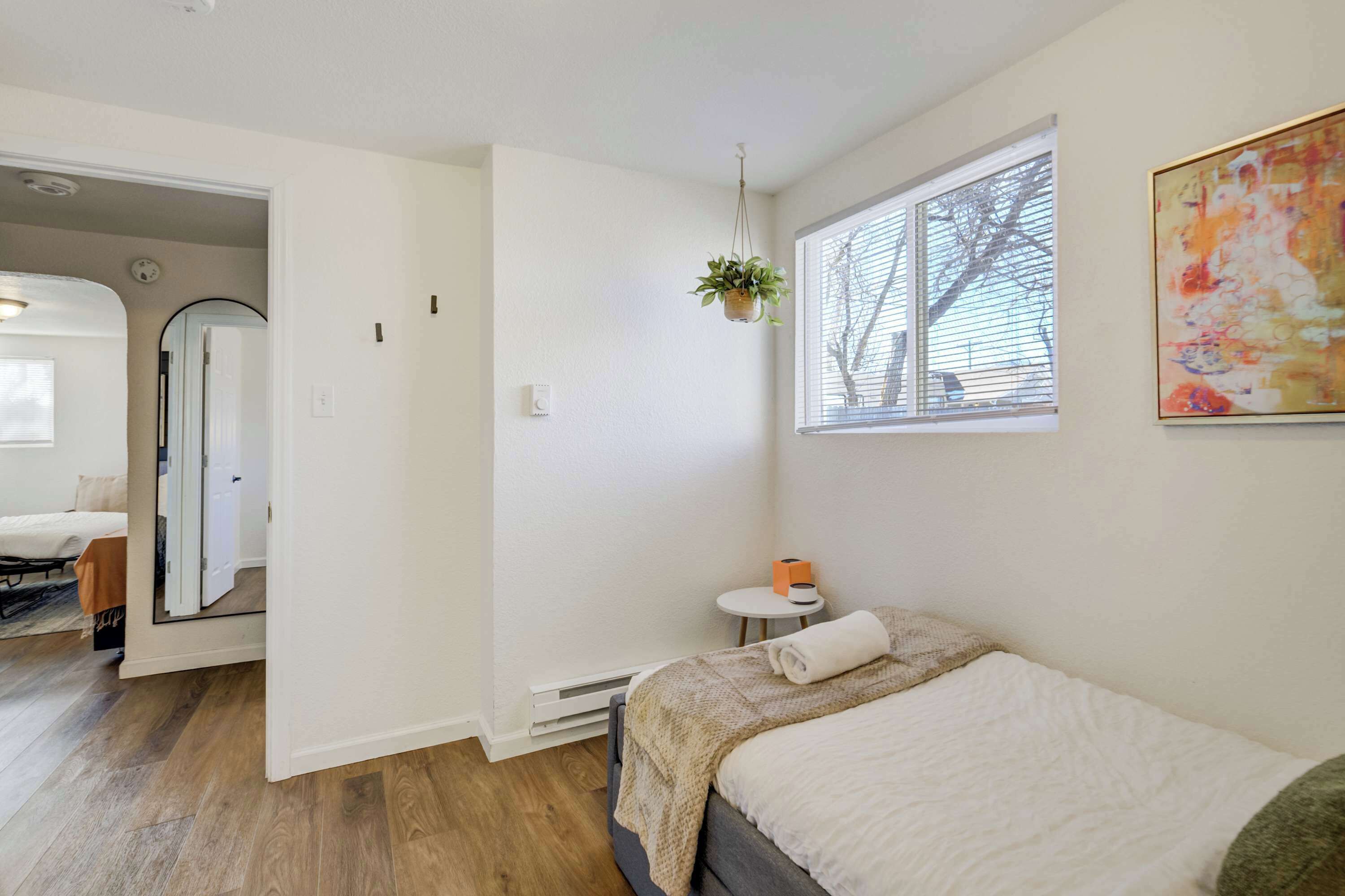A modern bedroom with a bed, a small bedside table, a hanging plant, and a window with blinds.