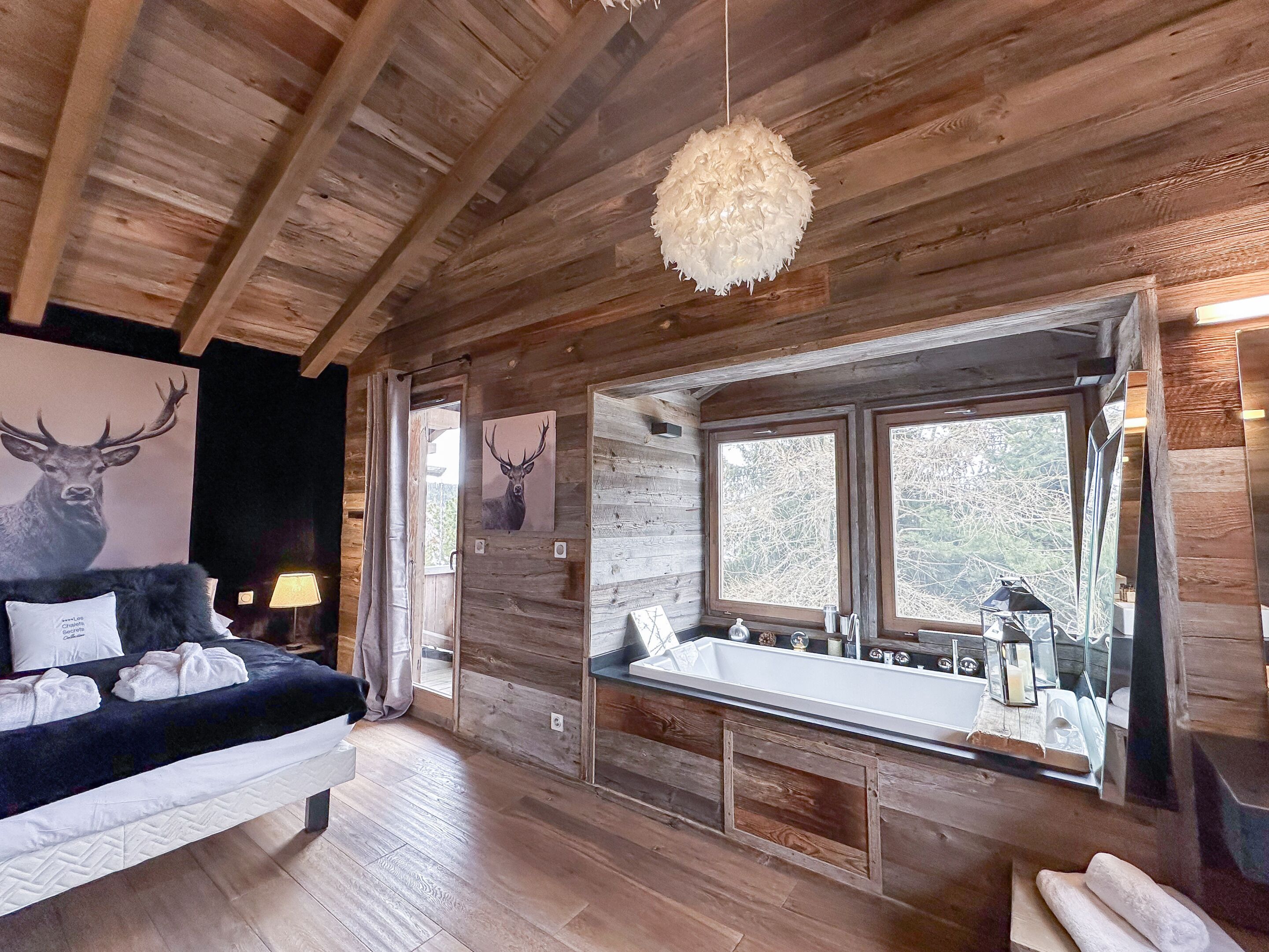 L’intérieur d’un chalet rustique, avec un plafond en bois, un lit recouvert d’une couette blanche et d’un couvre-lit foncé, une tête de cerf accrochée au mur, une fenêtre donnant sur les arbres et un miroir sur le mur.
