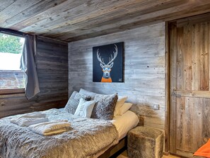 A rustic wooden room with a bed, a painting of a deer, and a window with a view of greenery.