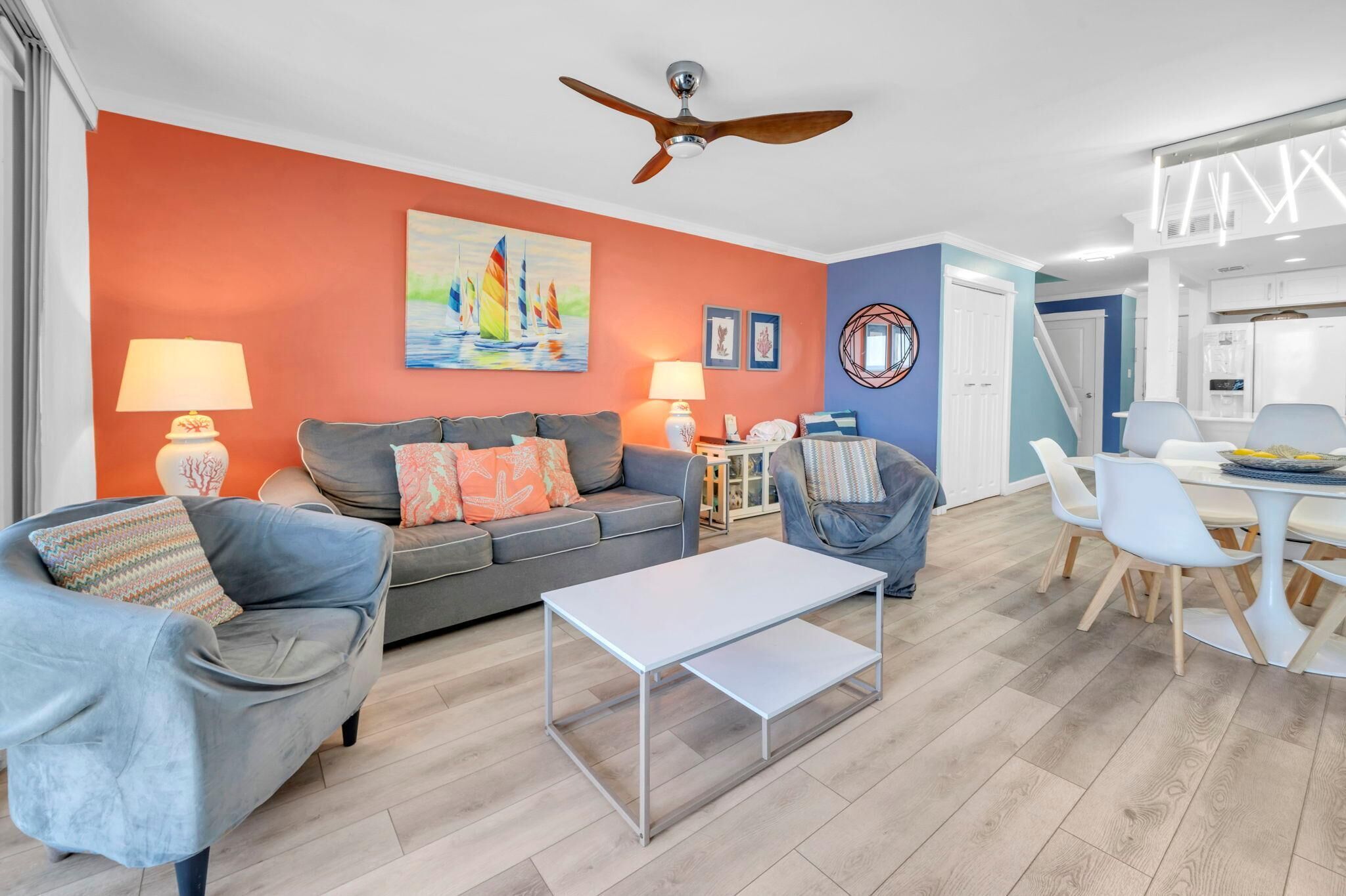 A modern living room with a grey sofa, a blue armchair, and a white coffee table. There is a dining area with a white table and chairs. The walls are painted in orange and blue, and there is a ceiling fan.