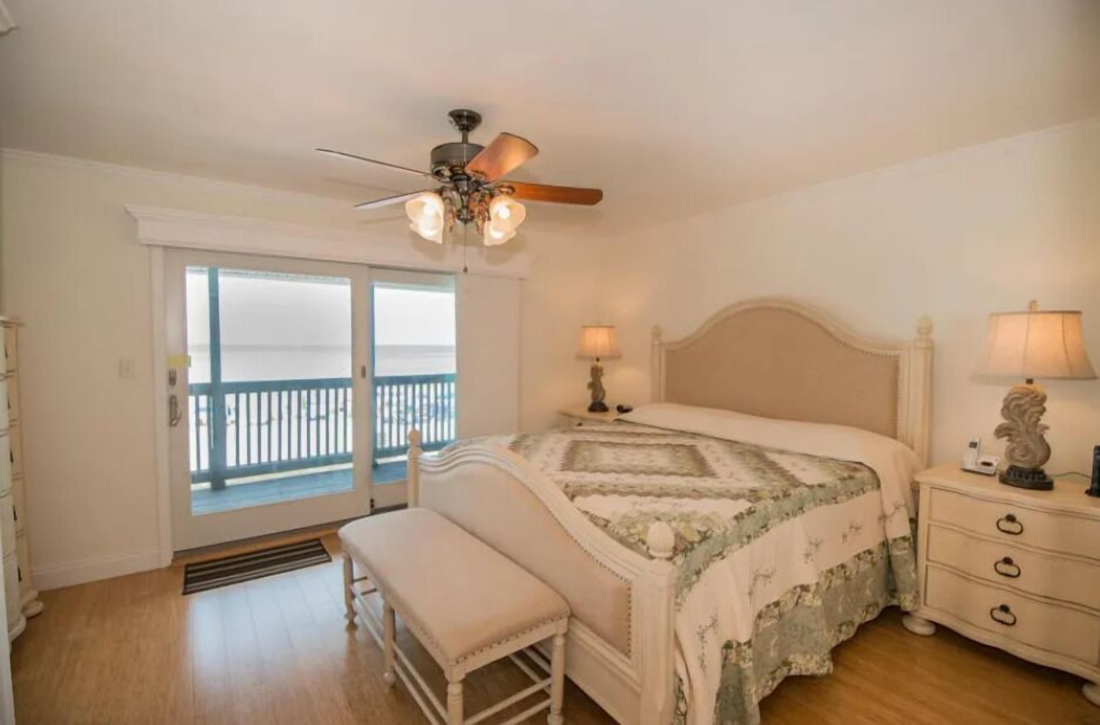 A bedroom with a bed, a bench, a nightstand, a ceiling fan, and a balcony with a railing.