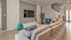 A modern living room with a sectional sofa, a flat-screen TV, a potted plant, and a staircase.