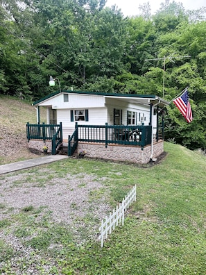 A white building with a green roof and a stone foundation, a flagpole with an American flag, and a white picket fence.