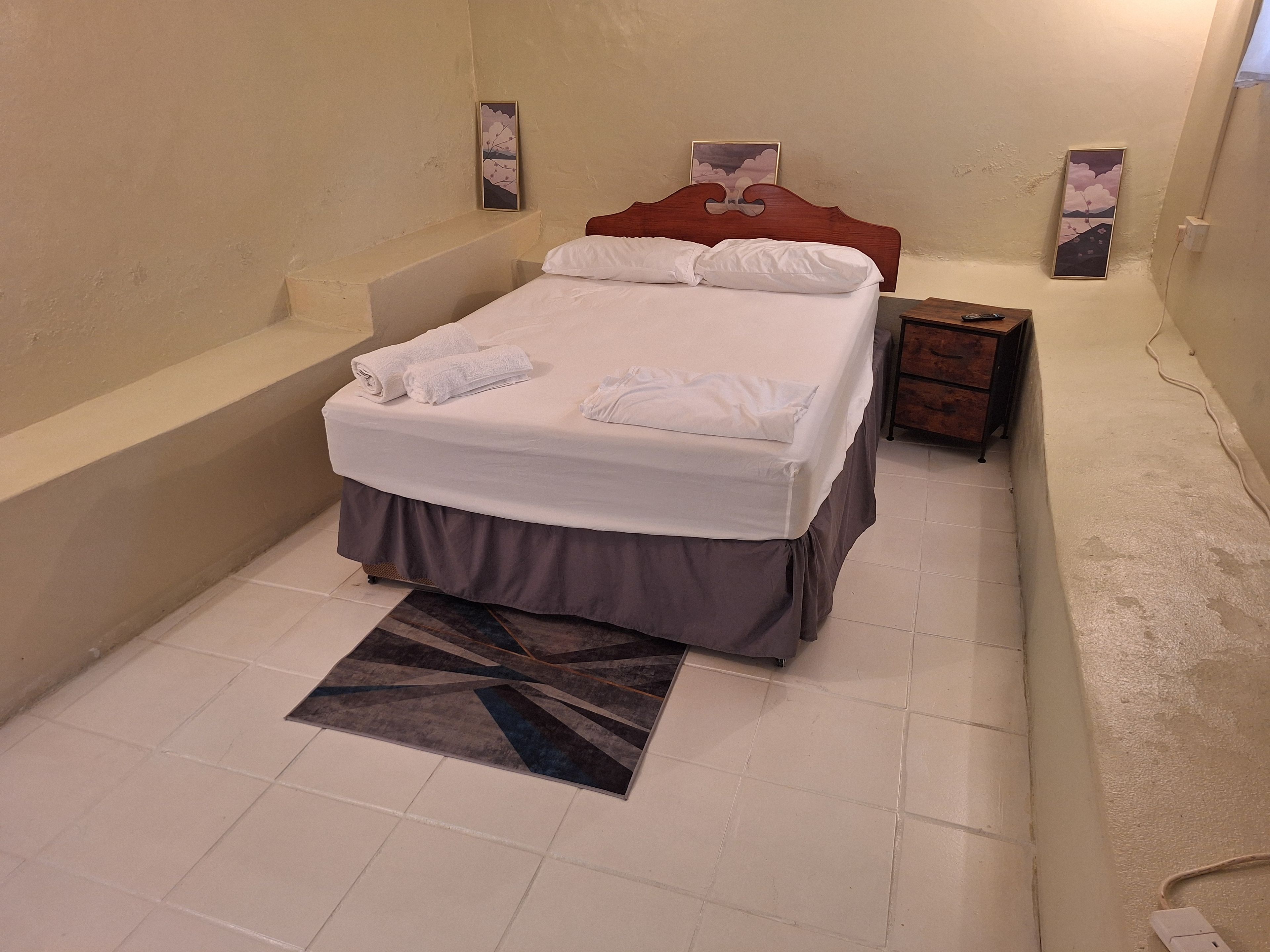 A bedroom with a bed, a nightstand, a wall-mounted artwork, and a tiled floor.