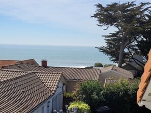 A coastal residential area with terracotta-tiled roofs, a large tree, and a view of the sea.