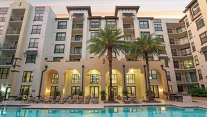 Ein mehrstöckiges Hotel mit Swimmingpool, Palmen und Liegestühlen.
