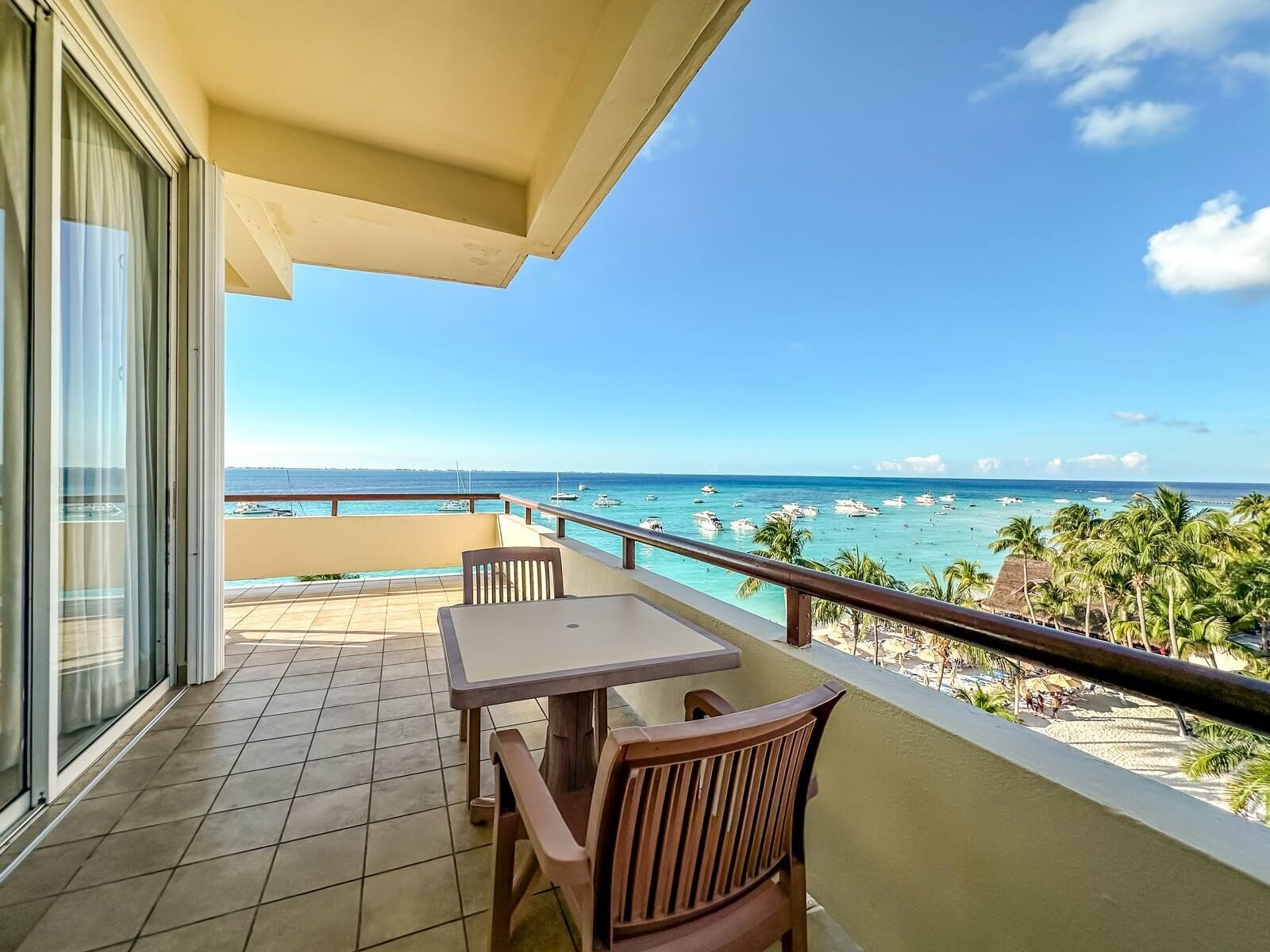 A balcony with a table and chairs, overlooking a beach with boats and palm trees.