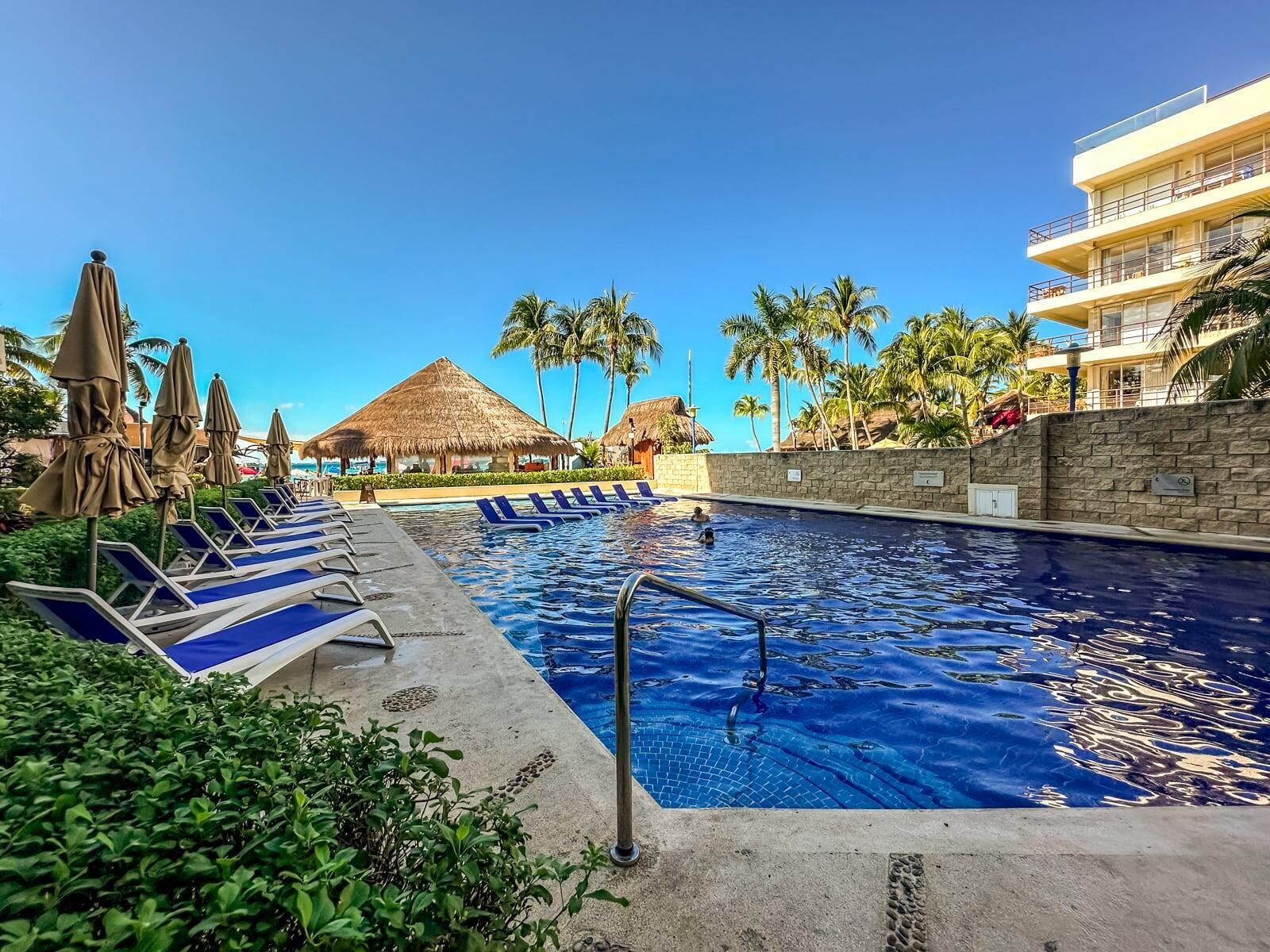A swimming pool with lounge chairs and umbrellas, surrounded by palm trees and a thatched-roof structure.