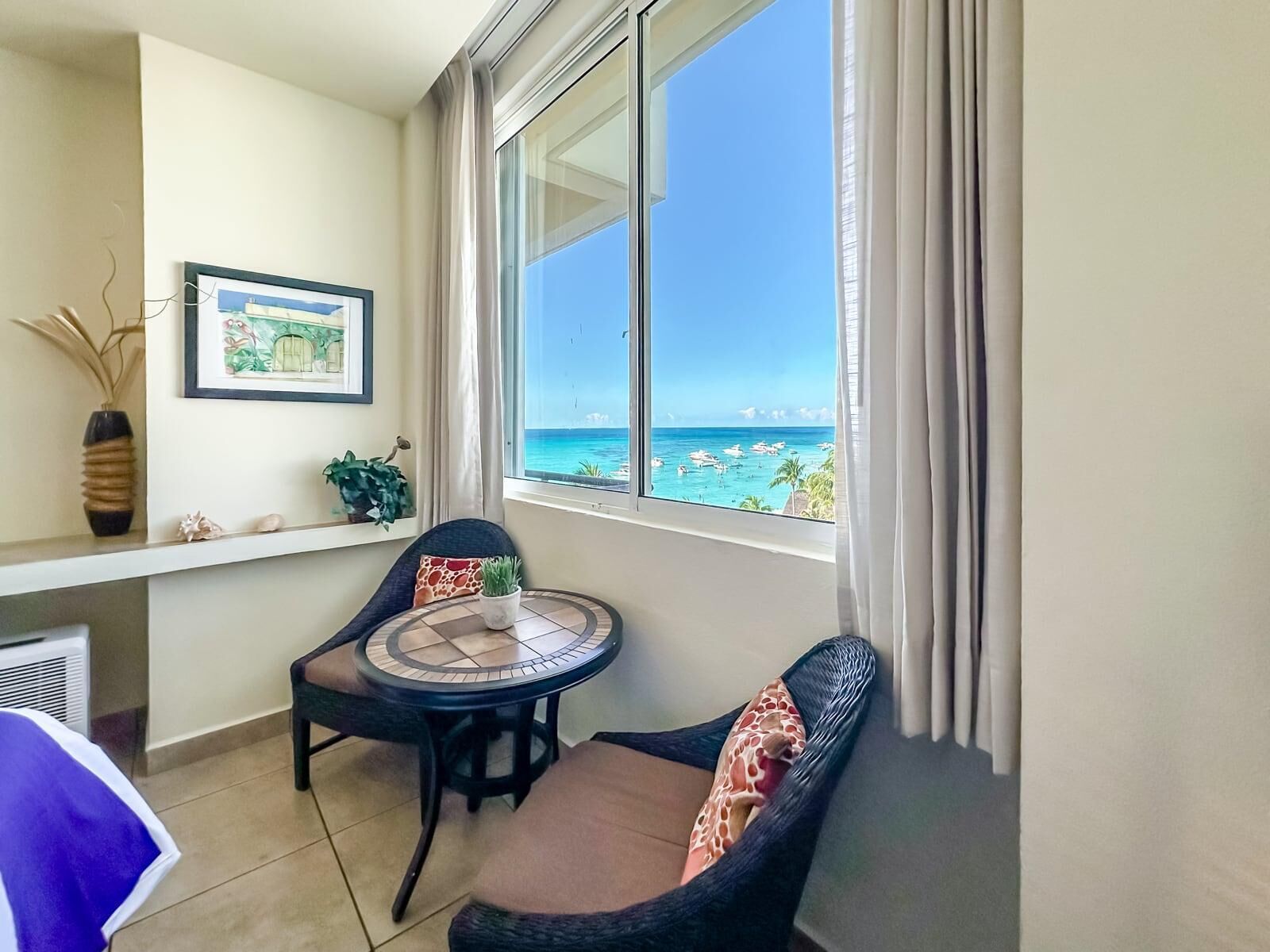 A balcony with a view of the sea, a chair, a small round table, and a framed picture on the wall.