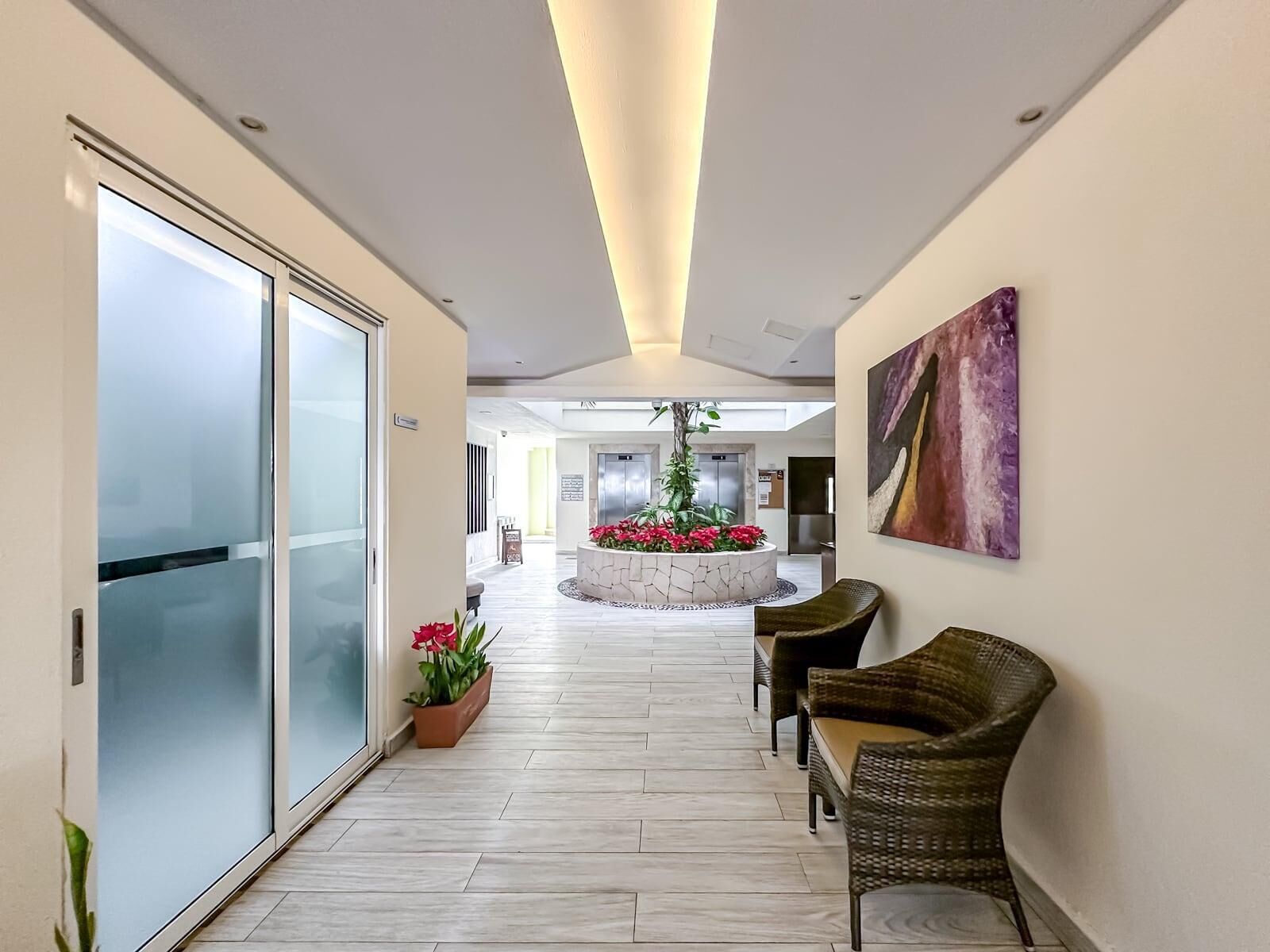 A modern hotel lobby with a glass door entrance, a central planter with flowers, and a seating area with wicker chairs.