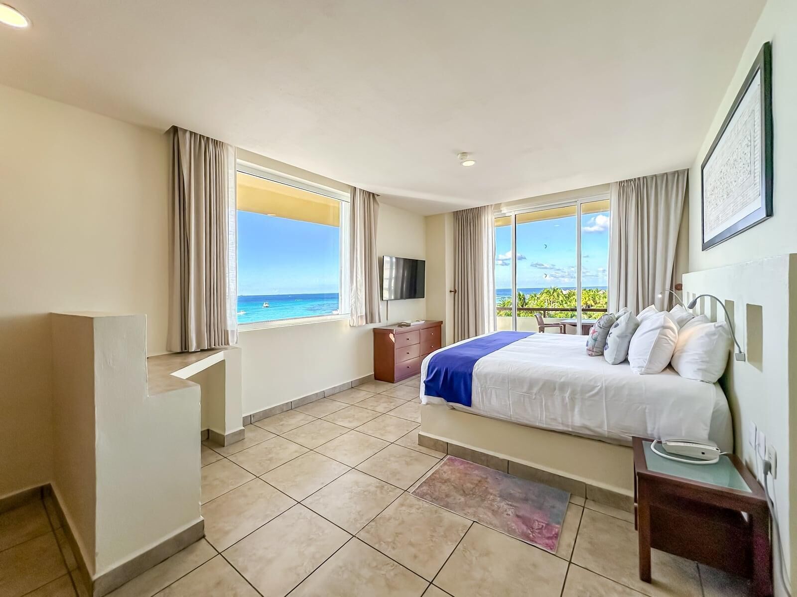 A hotel room with a bed, a TV, a desk, and a window with a view of the ocean.