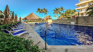 Une piscine entourée de palmiers et dotée de chaises longues et de parasols, ainsi qu’une structure à toit de chaume.