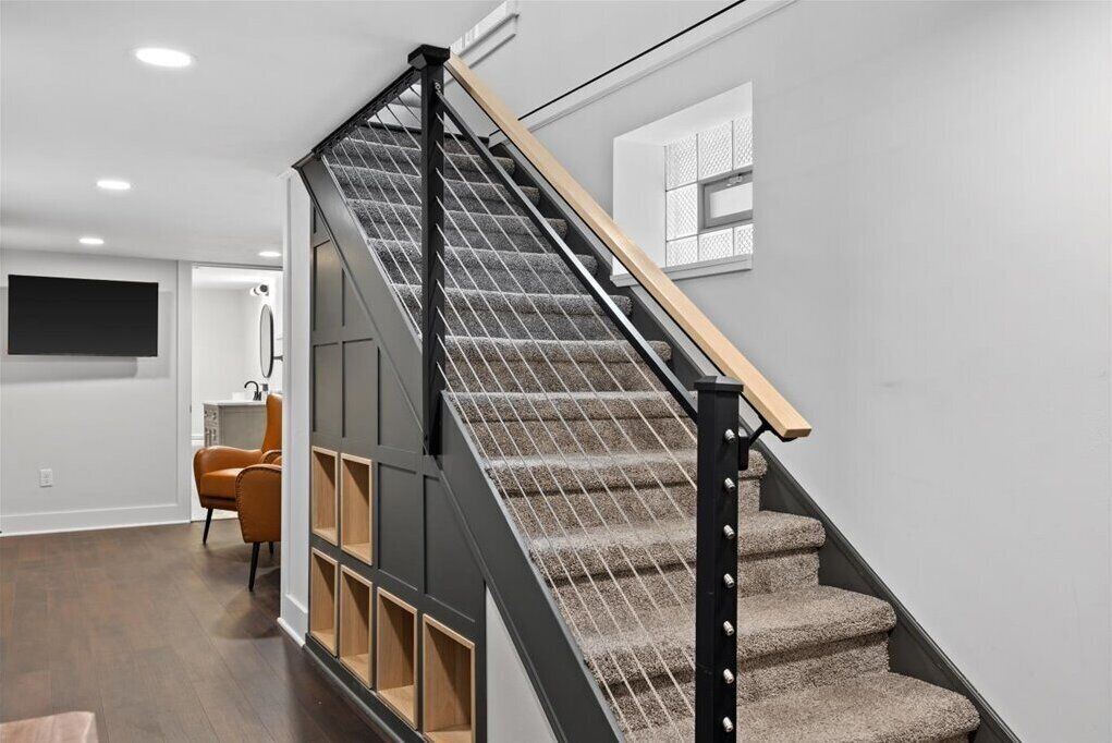 Un escalier moderne avec une rampe en bois et des marches recouvertes de moquette, menant à une pièce équipée d’un téléviseur à écran plat et d’un fauteuil.