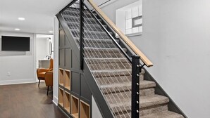 Un escalier moderne avec une rampe en bois et des marches recouvertes de moquette, menant à une pièce équipée d’un téléviseur à écran plat et d’un fauteuil.