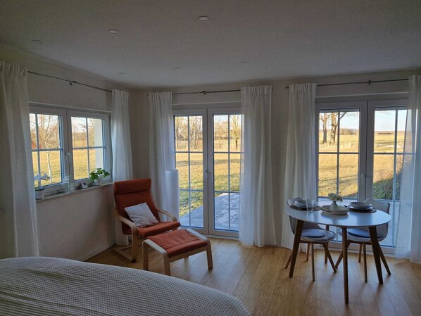 A room with a bed, a dining table with chairs, a red armchair, and large windows with curtains.