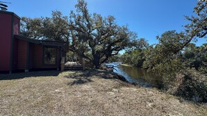 Una cabaña junto al río con un balcón, rodeada de árboles y un cielo despejado.