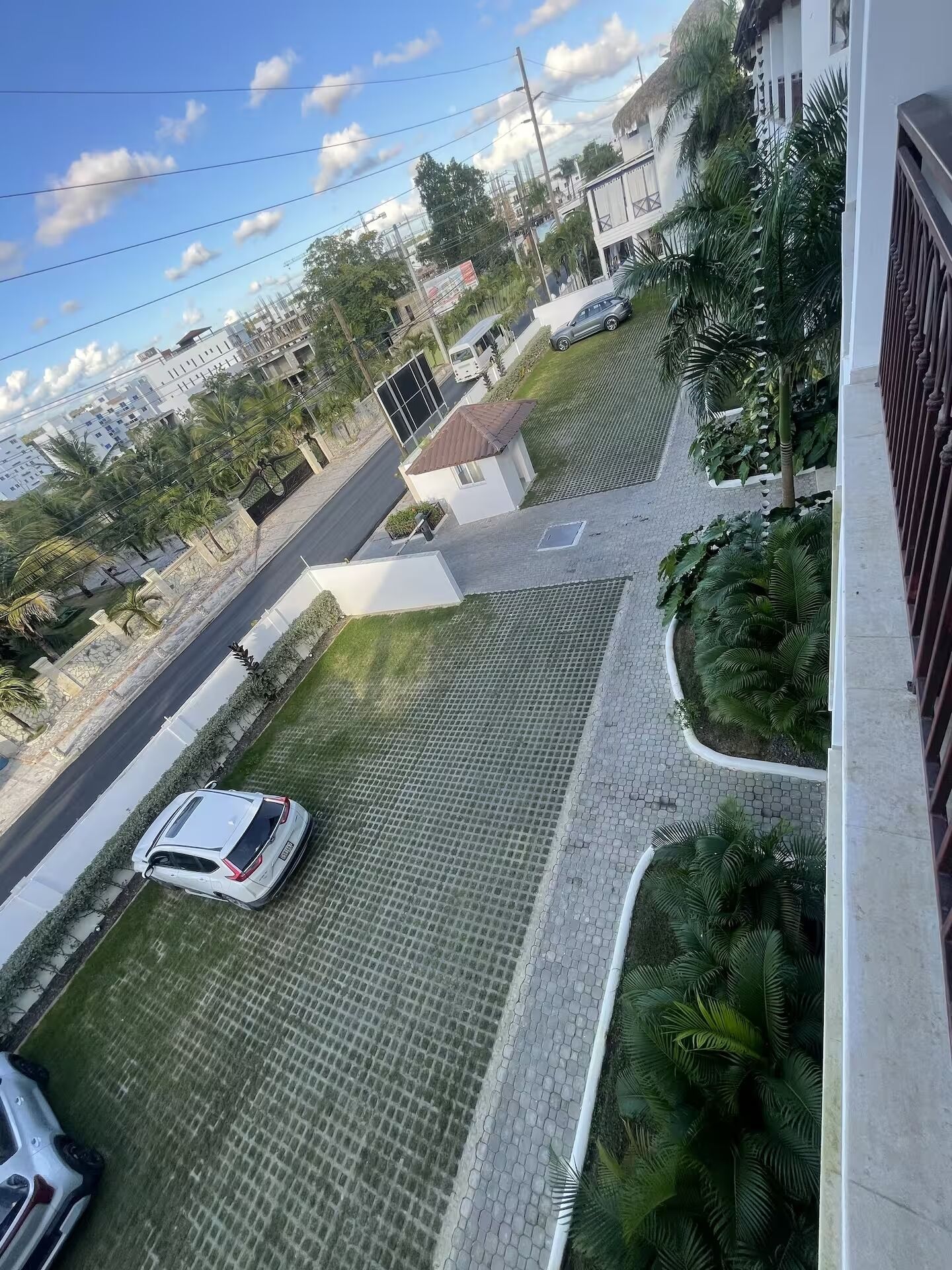 Zona residencial amb un carrer asfaltat, un cotxe blanc estacionat a la gespa, i un edifici amb teulada de palla.