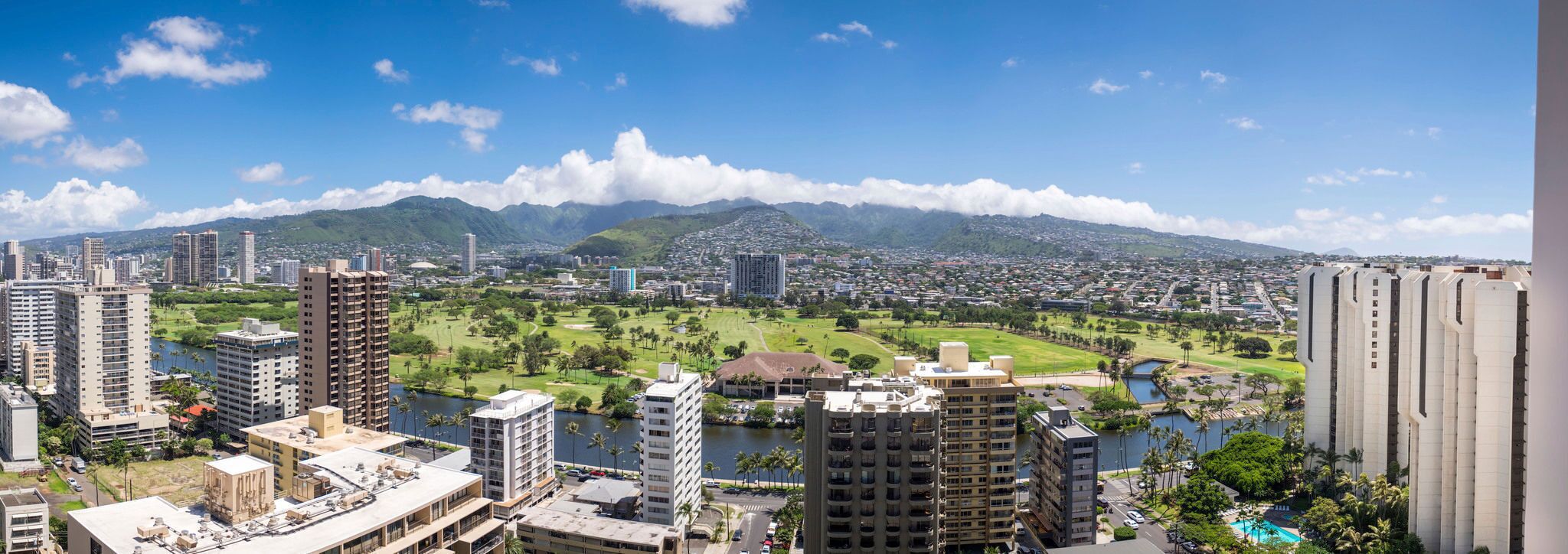 A cityscape with high-rise buildings, a golf course, and a mountain range in the background.