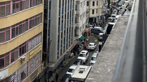 Une rue animée en milieu urbain, bordée d’immeubles à plusieurs étages, avec des voitures garées et une vue sur une étendue d’eau lointaine.