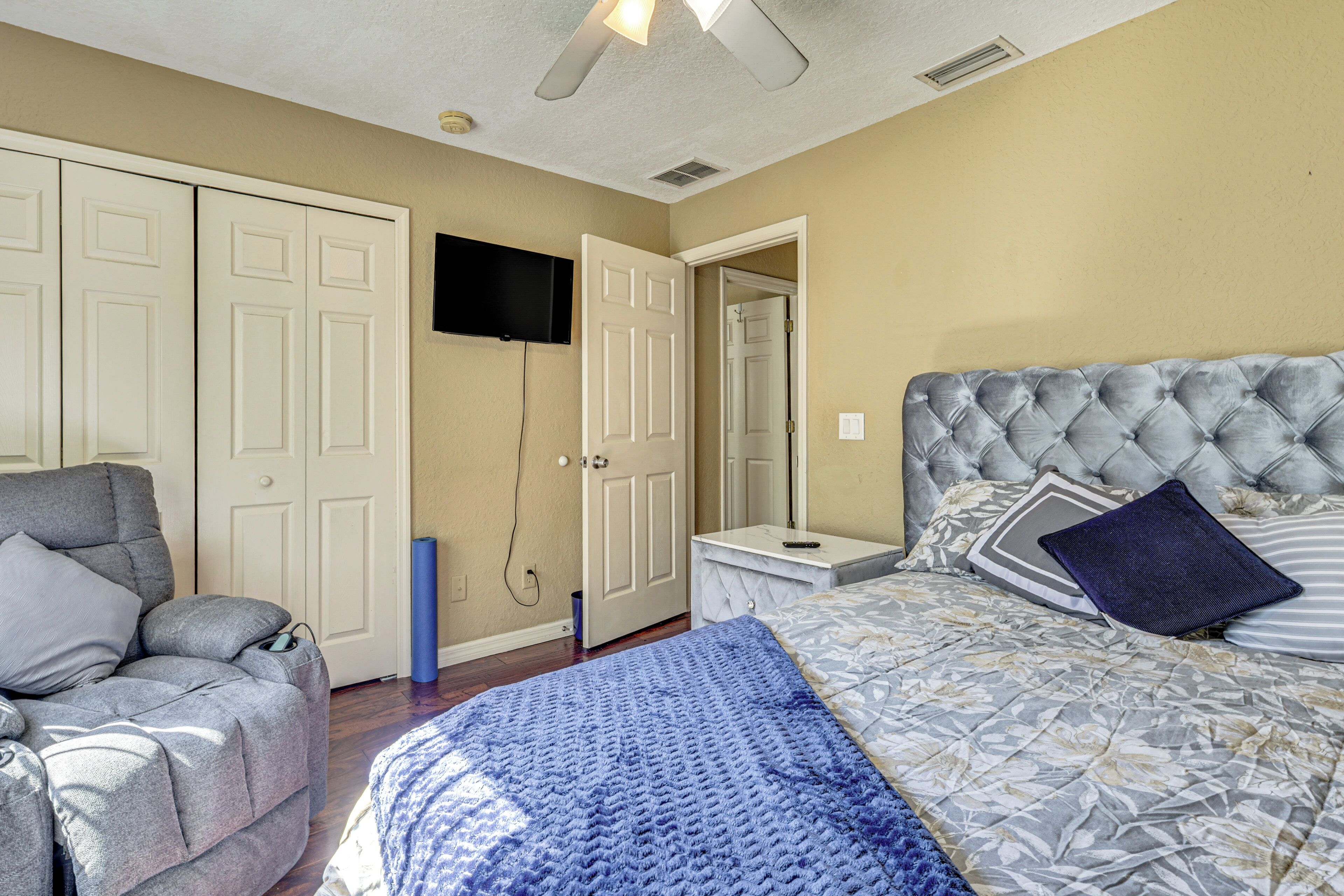 Un dormitorio con una cama, un sillón gris, un televisor montado en la pared y una puerta que da a otra habitación.