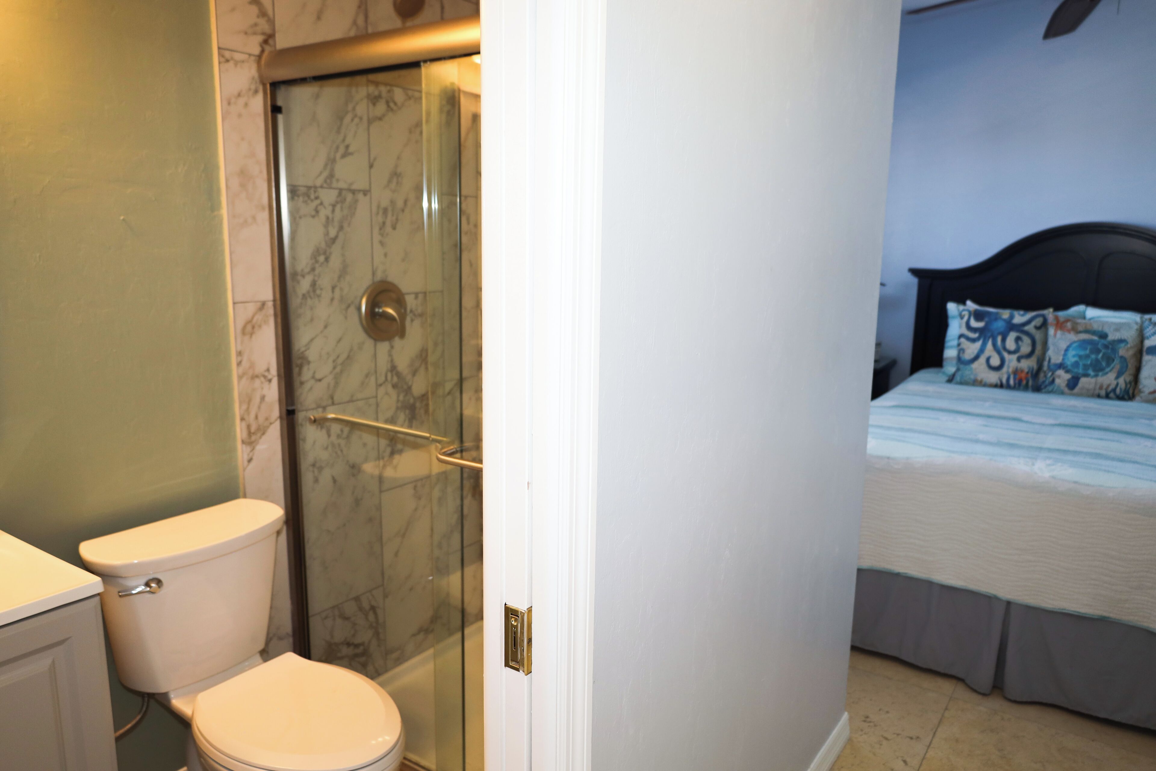 A bathroom with a toilet, a glass shower enclosure, and a bed with patterned pillows.