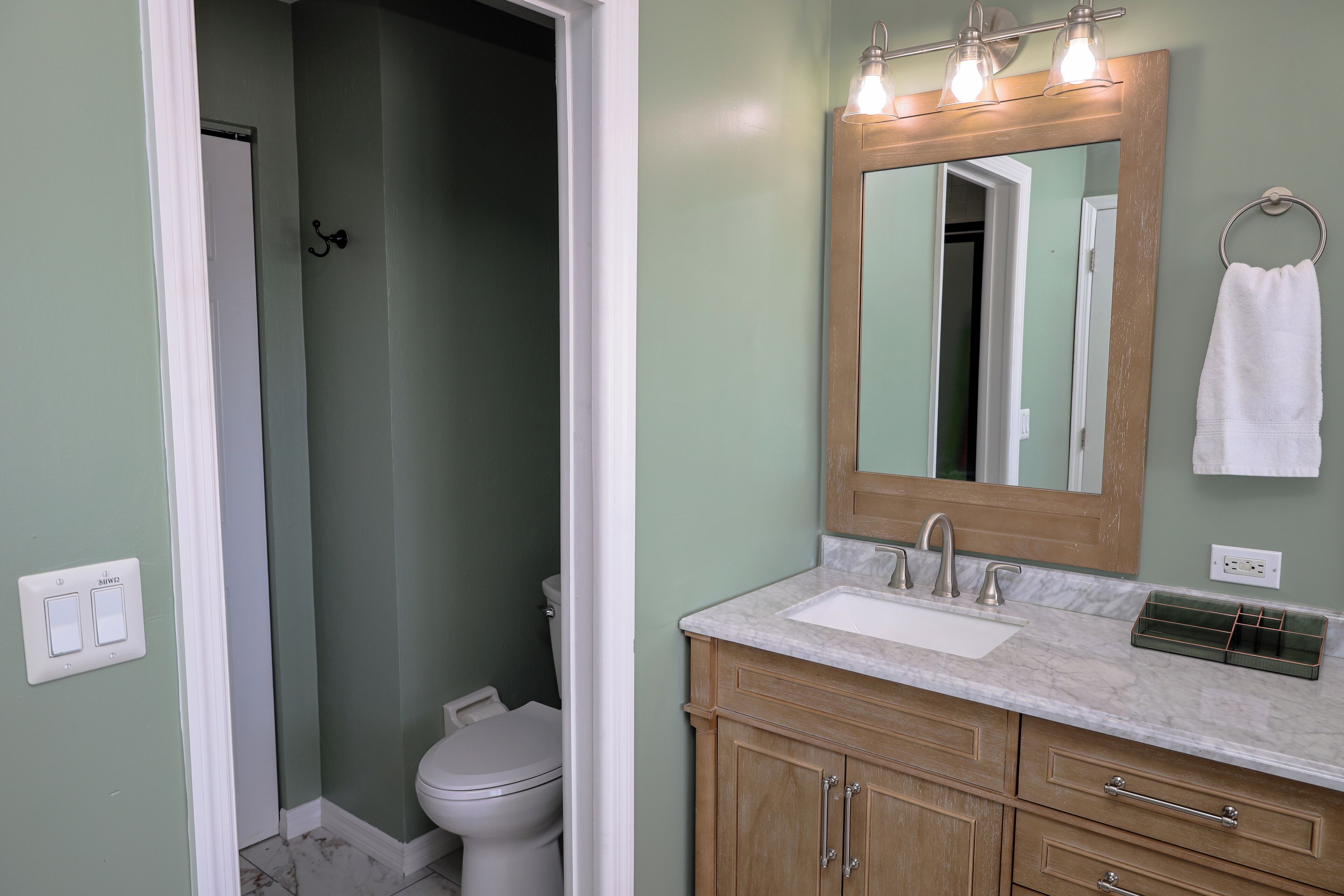 A bathroom with a white toilet, a sink with a marble countertop, wooden cabinets, and a mirror with lights above.