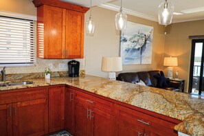 A kitchen with granite countertops, wooden cabinets, a coffee maker, and pendant lights. - Luxury Fort Myers Beachfront Penthouse - Sunset Views & Pool (Fort Myers Beach)