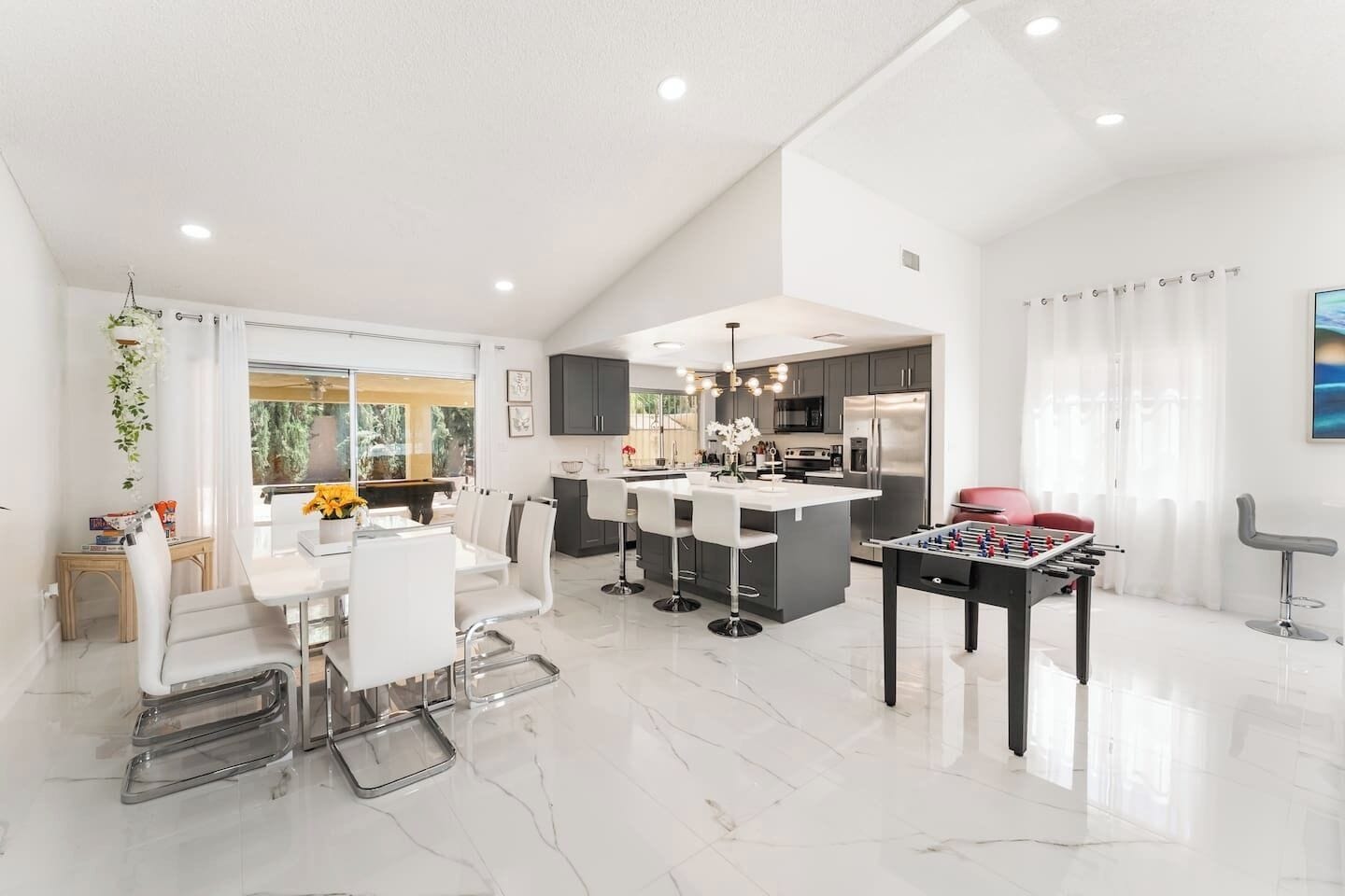 A modern kitchen with a central island, a dining area with a white table and chairs, and a foosball table.