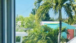 Une vue à travers une fenêtre donnant sur un quartier résidentiel, avec un petit arbre, une étendue d’eau et un bâtiment doté d’un toit bleu.