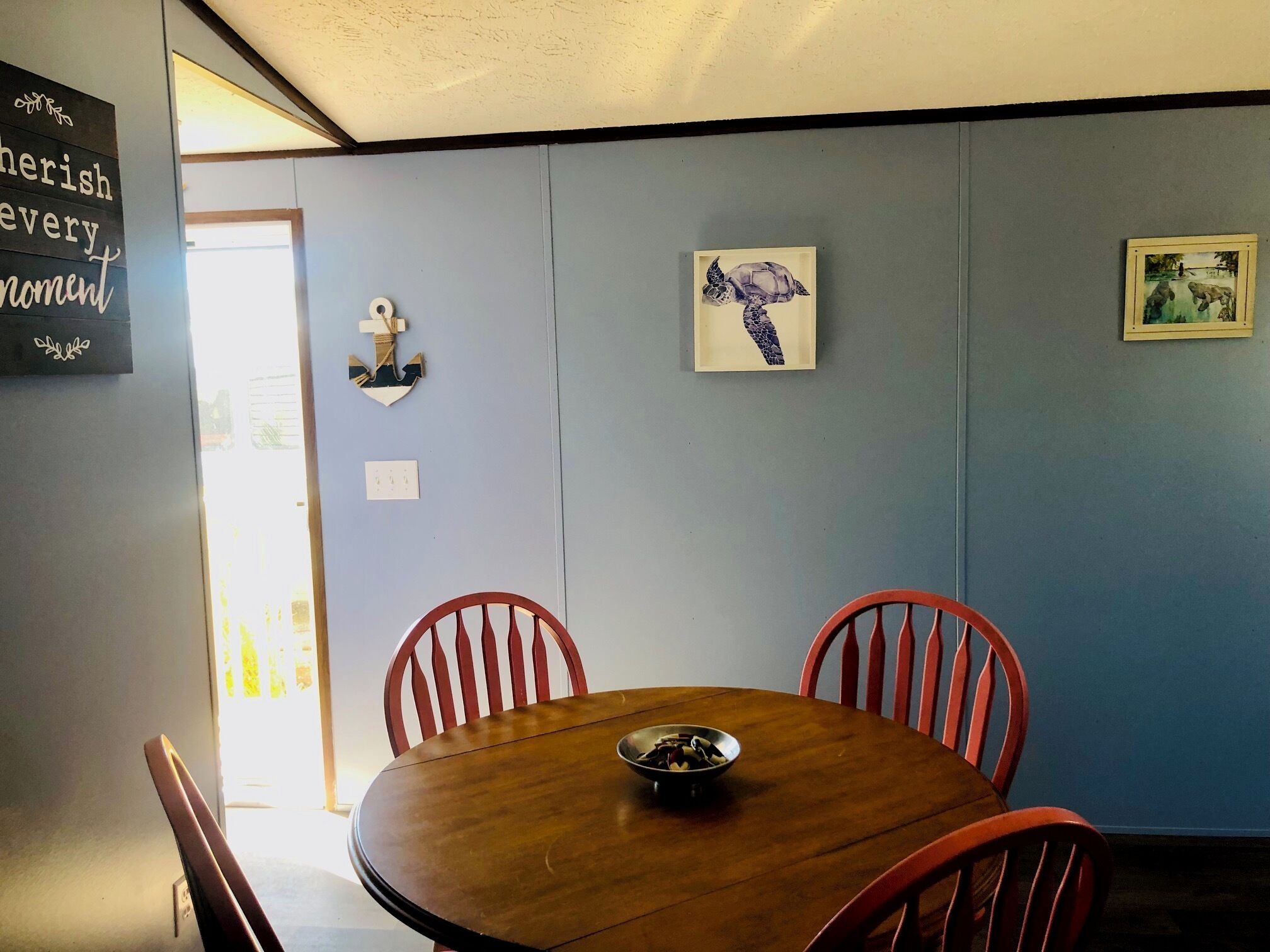 Un coin repas avec une table en bois et des chaises rouges, un tableau représentant une tortue encadré, et une décoration en forme d’ancre fixée au mur.