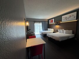Habitación de hotel con dos camas, un escritorio, una silla, una lámpara y un ventanal con cortinas.
