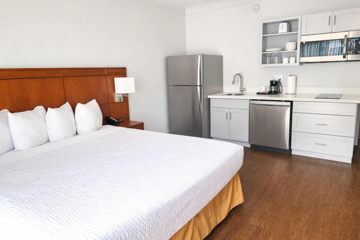 A hotel room with a large bed, a wooden headboard, a kitchenette with a refrigerator, microwave, and sink.