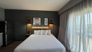Ein Hotelzimmer mit einem Bett, einem Kühlschrank, einem Nachttisch mit Lampe und einem fensterbelaubten Fenster.