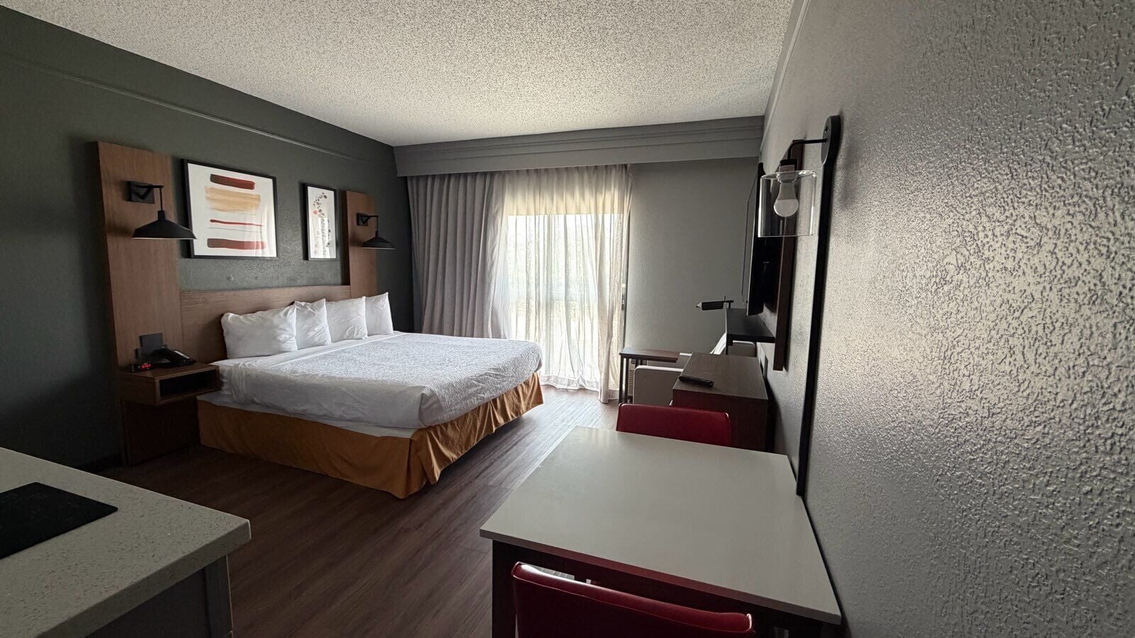 Une chambre d’hôtel comprenant un lit, une table de chevet, un bureau et une chaise, une fenêtre avec des rideaux et un téléviseur fixé au mur.