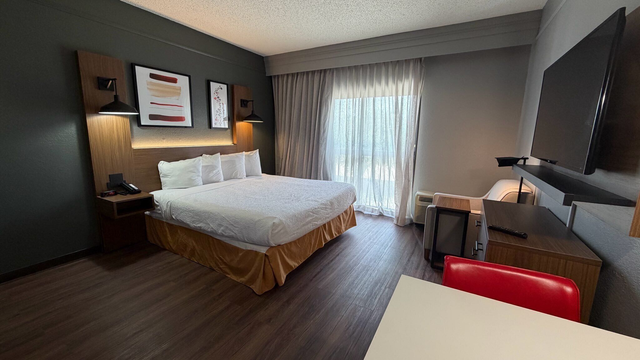 Une chambre d’hôtel comprenant un lit, une table de chevet, une lampe, des œuvres d’art, des rideaux et un téléviseur.