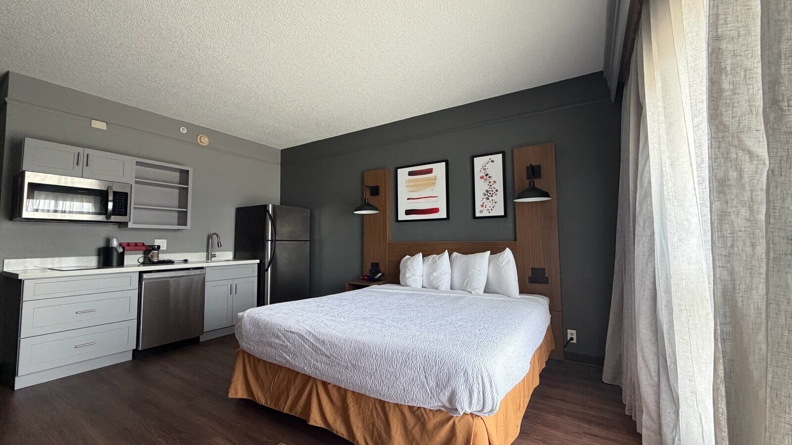 A compact hotel room with a kitchenette, a bed with a white duvet, and a wooden headboard.