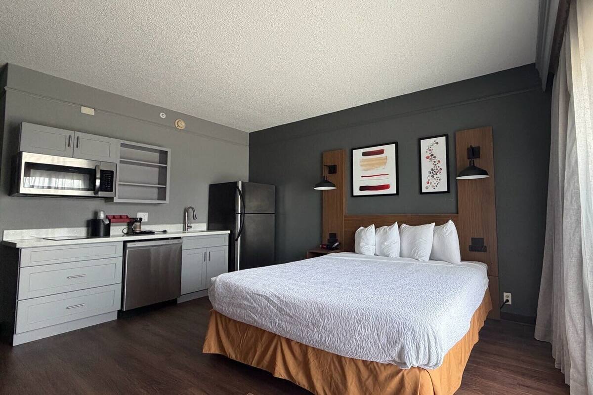 A compact hotel room with a kitchenette, a bed with a white duvet, and a wooden headboard.