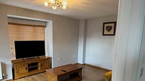 A living room with a flat-screen TV on a wooden stand, a coffee table, a sofa, and a wall-mounted light fixture.