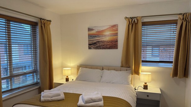 A hotel room with a bed, two bedside tables with lamps, a painting on the wall, and windows with curtains.